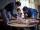 Une femme, un homme et un jeune garçon sont rassemblés autour d’une table circulaire. Plusieurs photographies sont éparpillées sur la table et la femme en montre une au garçon.